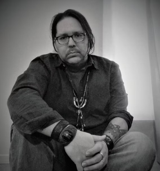 Black-and-white portrait of a man with glasses and long hair, seated with arms resting on knees, wearing a necklace and bracelets with tattoos showing on his forearm.