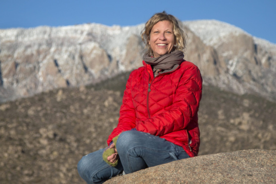 Woman in a red jacket sits on a rock with snow-capped mountains in the background, smiling at the camera.