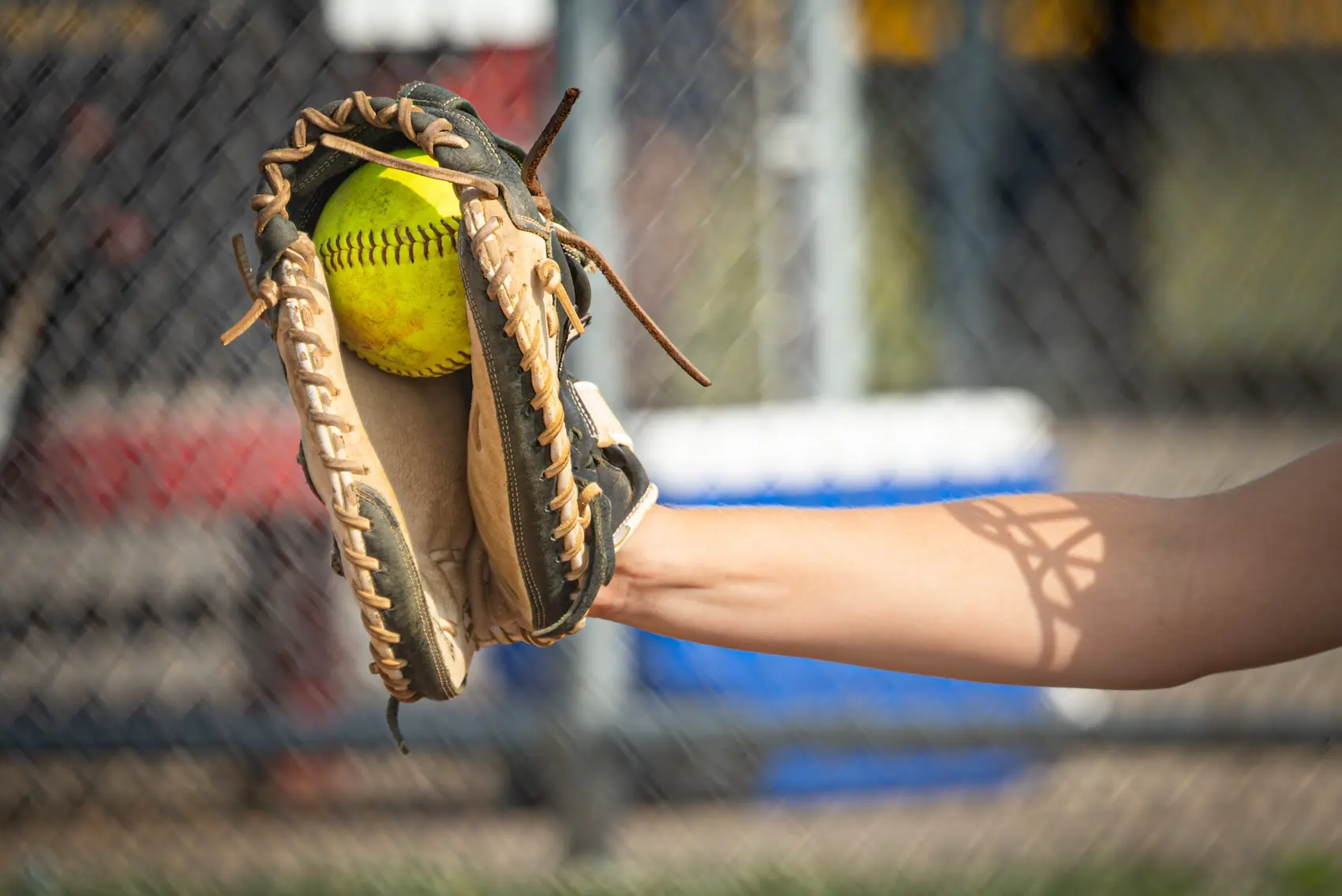 A worn baseball glove catching a yellow softball against a blurred chain-link fence backdrop, held by an outstretched arm.