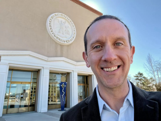 Smiling man in a suit taking a selfie outside a beige building with a round seal emblem above the entrance.