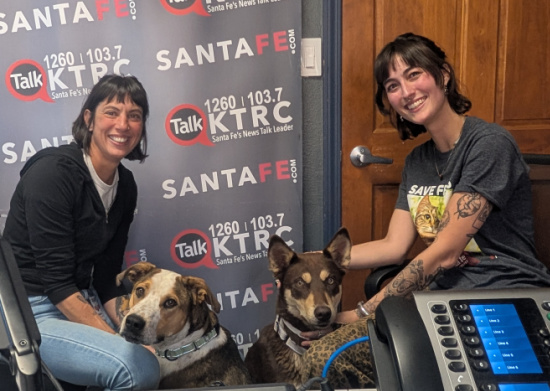 Two smiling women sit with two dogs inside a radio station booth; backdrop reads Santa Fe Talk 1260/KTRC and SantaFe.