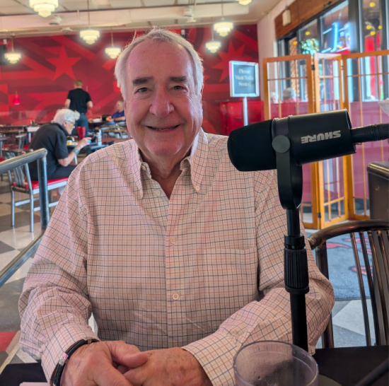 Smiling older man in a light plaid shirt sits at a cafe table with a Shure microphone in front, as if recording a podcast.