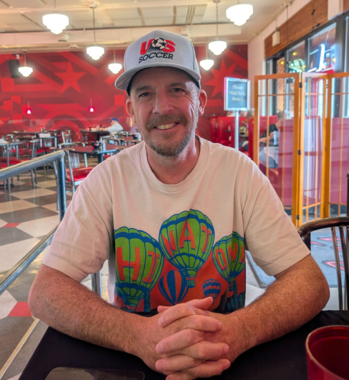 Smiling man wearing a gray USA Soccer cap and a light T‑shirt with a hot air balloon design sits at a table in a bright, retro-style cafe.