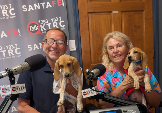 Two radio hosts sit at microphones in a studio, each holding a small dog, with station banners in the background.