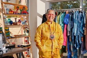 Smiling person in a yellow paint-splattered jumpsuit stands among colorful clothing on racks and handmade items on shelves in a boutique storefront.