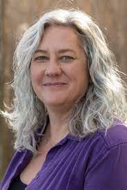 Portrait of a smiling woman with gray curly hair wearing a purple blouse outdoors.
