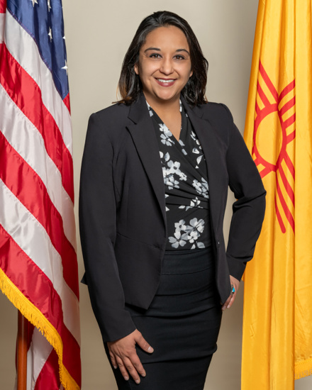Portrait of a woman in a dark blazer standing between the American flag and a yellow flag, smiling at the camera.