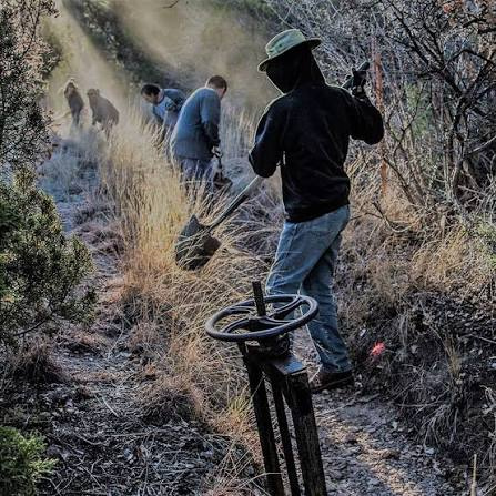 Person wearing a black hoodie and mask uses a metal detector on a dry, scrubby hillside while others dig in the background; a search or artifact-hunt scene.