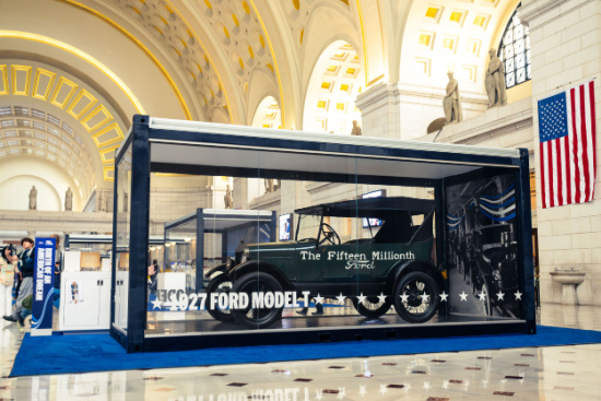 In a grand museum hall, a glass display case houses a 1927 Ford Model T labeled 'The Fifteen Millionth' with an American flag in the background.