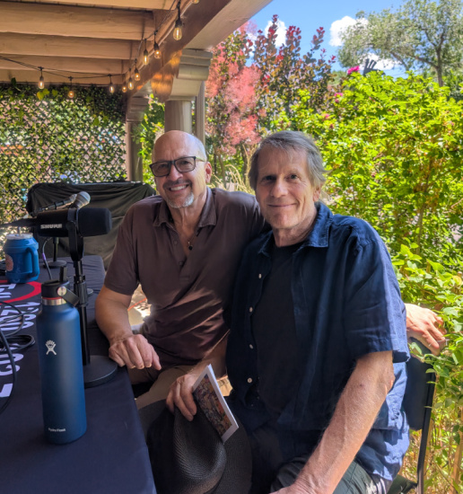 Two men sit at a patio table with a microphone setup, smiling for the camera in a sunny outdoor space with greenery around them.