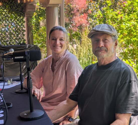 Two hosts sit at a table with a microphone outdoors under a pergola; a smiling woman in a pink blouse and a man in a cap pose for the camera.
