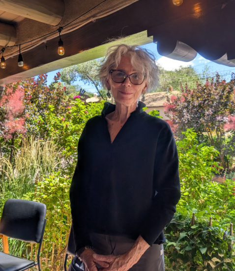 Older woman with gray hair and glasses standing outdoors in a garden, wearing a black sweater under a canopy with string lights overhead.