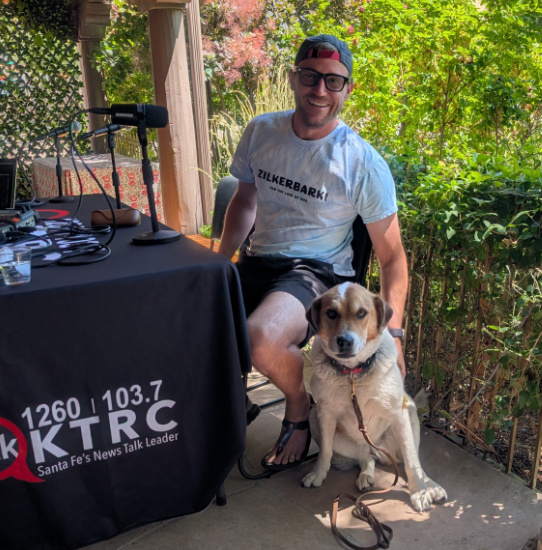 Man in a cap and glasses sits at a table with a microphone outdoors, smiling, with a dog sitting beside him.
