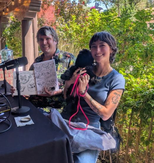 Two people sit at a table outdoors with a microphone; the man shows a sketchbook while the woman holds a small black dog with a pink leash.