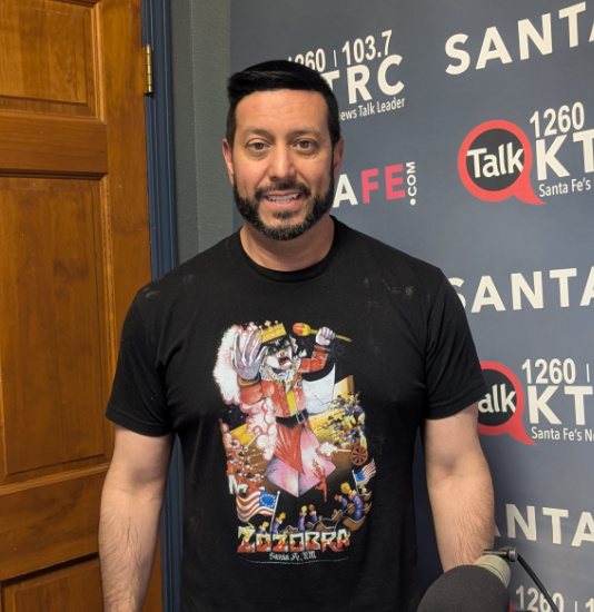 Man with dark hair and beard wearing a black graphic T-shirt, standing in a radio studio with a blue backdrop and station logos nearby, microphone in front.