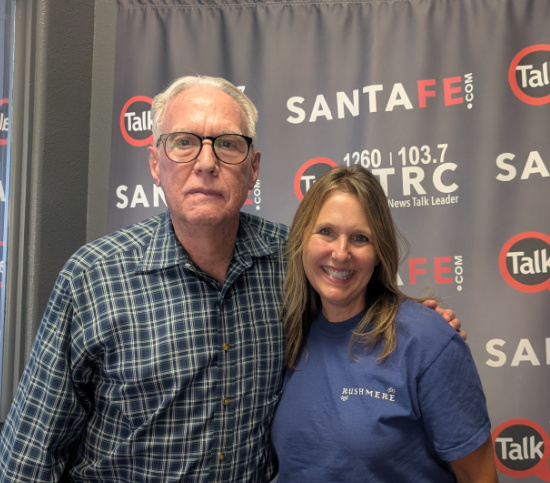Man in a plaid shirt and glasses and a smiling woman in a blue Rushmore shirt pose together in front of a Santa Fe radio backdrop.