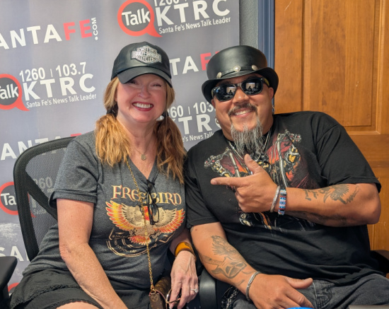 Two people smiling in a radio studio in front of a KTRC Santa Fe backdrop; woman wears a gray tee and cap, man wears a black graphic shirt with sunglasses and tattoos in view.