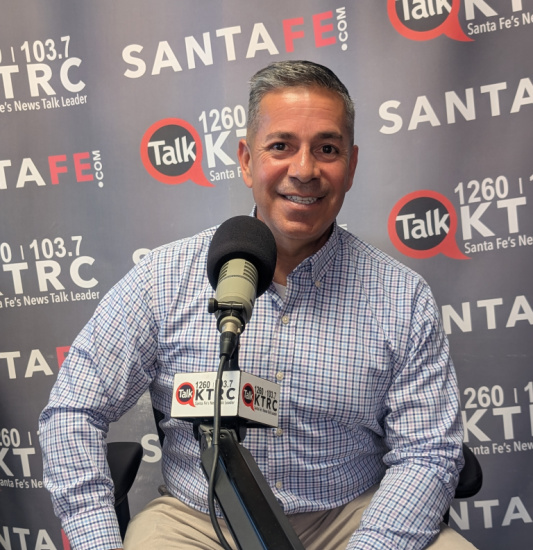 Smiling man in a blue checkered shirt sits at a radio studio microphone, with a branded backdrop reading Talk 1260 KTKC and Santa Fe logos on the wall.