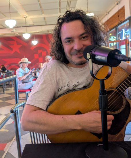 Musician with curly hair plays an acoustic guitar in a cafe, singing into a Shure microphone.