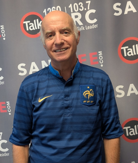 Smiling older man in a blue French football federation jersey stands in front of a backdrop with Talk Leader logos.