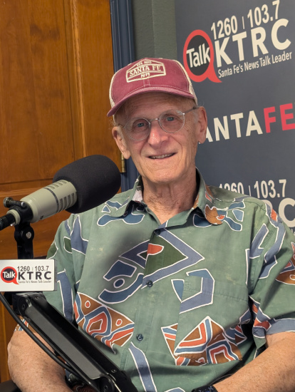Older man wearing a maroon cap and teal patterned shirt sits in a radio studio, speaking into a microphone with a KTCR banner behind him