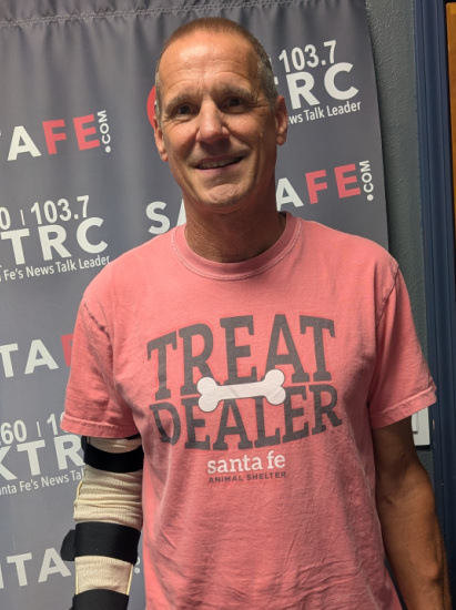 Smiling man in a pink shirt that reads 'TREAT DEALER' with a wrapped arm, standing in front of a radio-station backdrop.