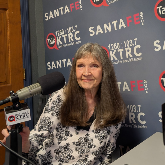 Smiling woman with long gray hair, wearing a white and black patterned blouse, sits at a radio desk with a microphone in front of a KTCR backdrop.