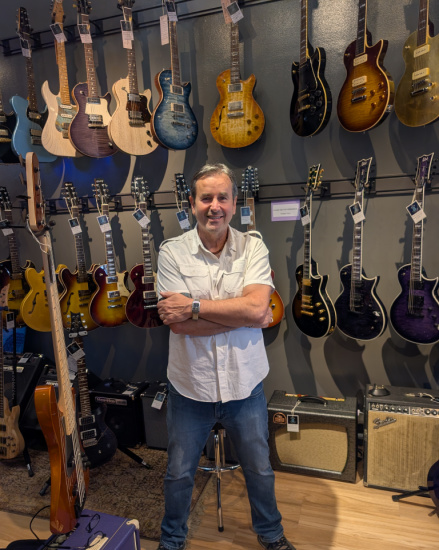 Man stands with arms crossed in a guitar store showroom, surrounded by rows of electric guitars hanging on the wall and amplifiers on the floor.