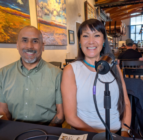 Smiling man in a green shirt and smiling woman in a white top sit at a cafe table; a microphone on a stand sits in front of the woman, suggesting a podcast recording.