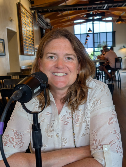 Smiling woman seated at a table with a microphone in a sunlit cafe-like recording space, ready to record a podcast.