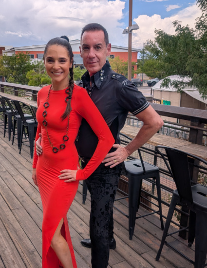 Smiling couple dressed for dance pose on an outdoor wooden deck; woman in a red dress with a slit, man in a black shirt beside her.
