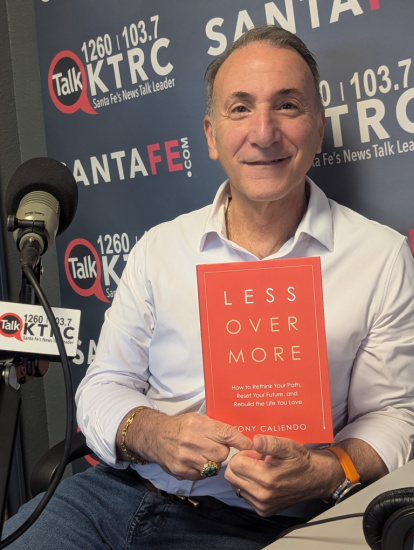 Man in a radio studio holds a red book titled 'Less Over More' during an interview, with Talk KTRC banners in the background.