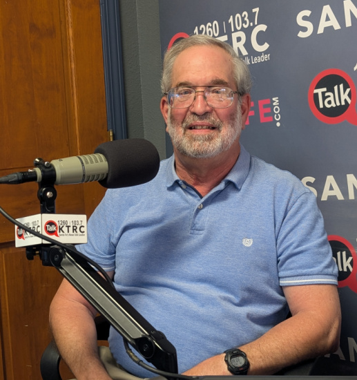 Man in a light blue polo shirt sits in a radio studio in front of a large mic, with a banner backdrop.