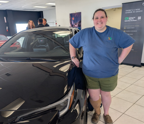 Smiling woman in a blue shirt and green shorts stands beside a black car inside a car dealership showroom.