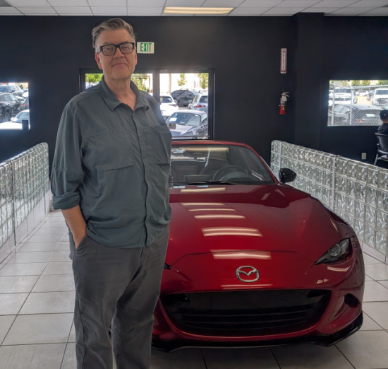 Man in a blue-gray shirt and glasses stands with hands in pockets beside a red Mazda convertible in a showroom