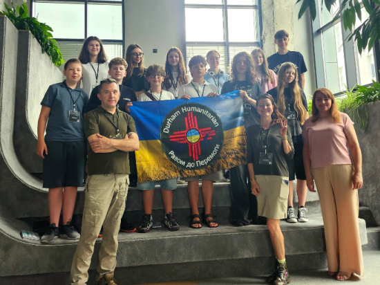 Group of about 15 young people and two adults on a stairwell indoors, holding a blue-yellow flag with a red and white circular humanitarian logo in the center.