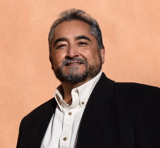 Portrait of a smiling man with salt-and-pepper hair and beard, wearing a white shirt and black blazer, against a peach wall.