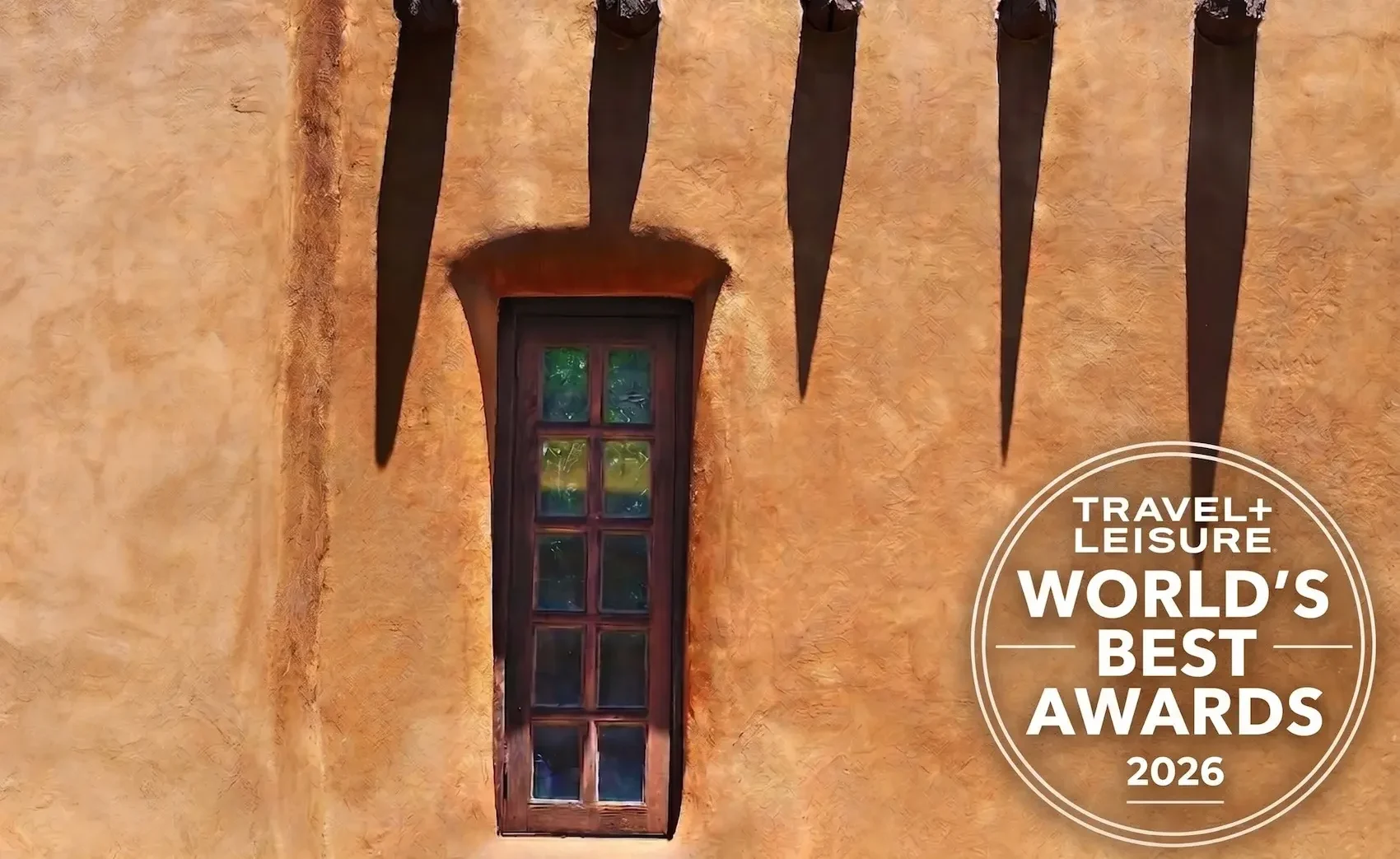 Orange stucco wall with a narrow wooden window; five tall dark shadows rise from the top, plus a Travel + Leisure World's Best Awards 2026 badge on the bottom-right.