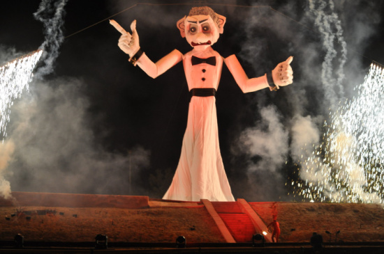 Giant stage puppet in a pink gown and black bow tie, arms raised, with pyrotechnics and smoke around.